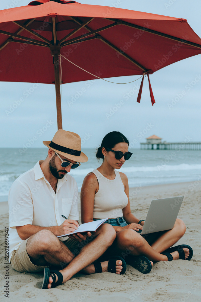 Fototapeta premium Vertical image of Caucasian young adult couple working on beach under umbrella using laptop and writing in notebook, representing remote work lifestyle and digital nomad career concepts