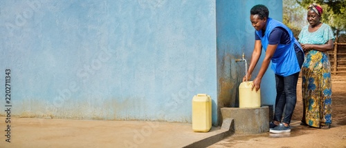 In a picturesque village setting during daylight hours, two individuals are busily filling their containers with water from a nearby water source