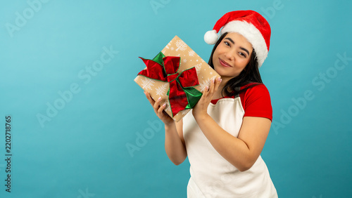 Young latina woman wearing santa hat holding a christmas present on a blue background