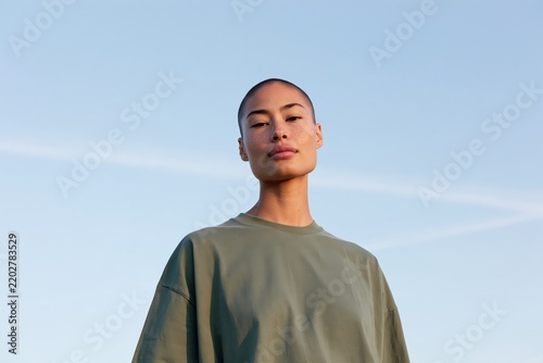Minimal outdoor portrait of a young woman with shaved head in a green oversized tee against a clear blue sky for lifestyle background poster and banner design