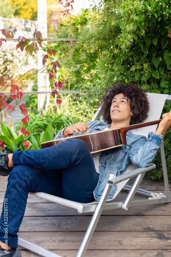 Beautiful young Afro-Italian mixed race man in casual wear playing guitar in the garden. A multiracial boy plays a musical instrument rests and relaxes outdoors. Spending time at home