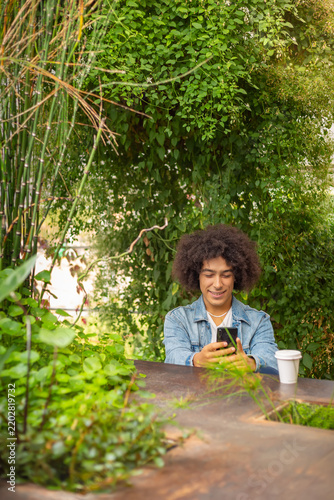 Smiling young ethnic guy 20 years old, dressed in casual clothes, works on a laptop outdoors, in a green area in nature. Green open space for coworking, freelancer, relaxing, coffee, food. Free place
