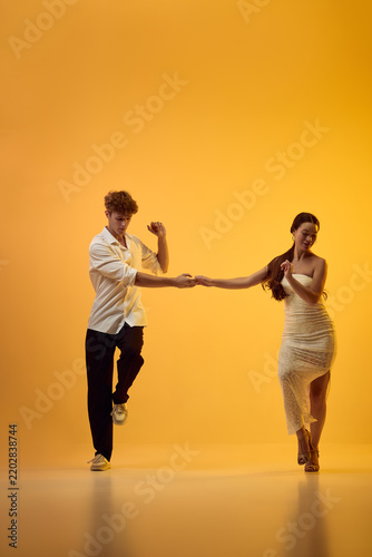 Bachata partners hold hands while stepping apart in orange studio with playful anticipation. Social dancing, couple interaction and rhythmic connection concept.