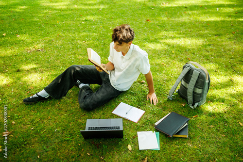Focused caucasian teenager reading a book while sitting on the campus grass.