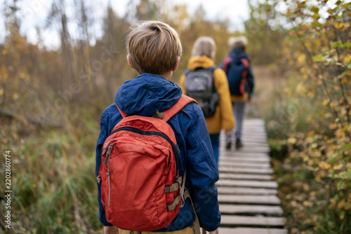School children crossing small wooden bridge during autumn hiking trip, focus on one child with backpack, teamwork and outdoor adventure concept.