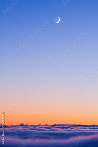 Crescent moon shining in a vibrant twilight sky above a thick layer of soft rolling clouds