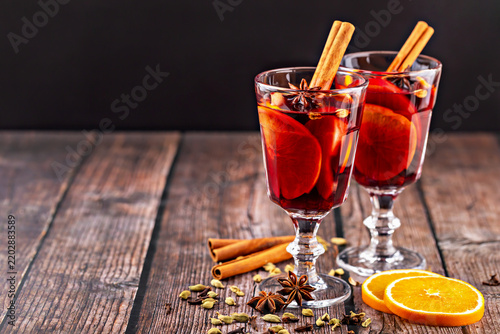 Two glasses of aromatic mulled wine on clear stems with orange slices and cinnamon sticks, while citrus rounds, star anise, cardamom, and extra cinnamon rest on wooden table