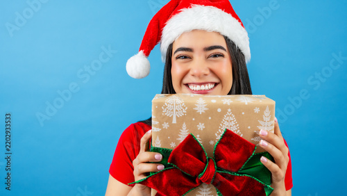 Smiling Latina woman wearing a santa hat and red shirt holding a wrapped gift box