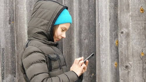 Small beautiful girl in hood and jacket looks at phone against tall wooden fence in autumn.
