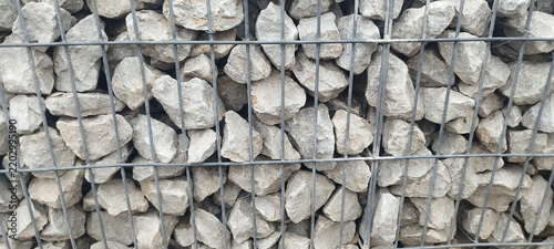 Фотография Close-up shot of a metal wire mesh gabion cage filled with rough grey crushed limestone rocks, forming a sturdy textured retaining wall pattern in bright daylight