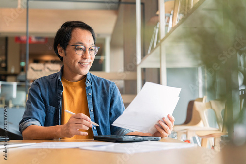 Focused Asian consultant reviewing business paperwork with content expression, analyzing financial report and strategic contract while planning project development in modern bright creative office