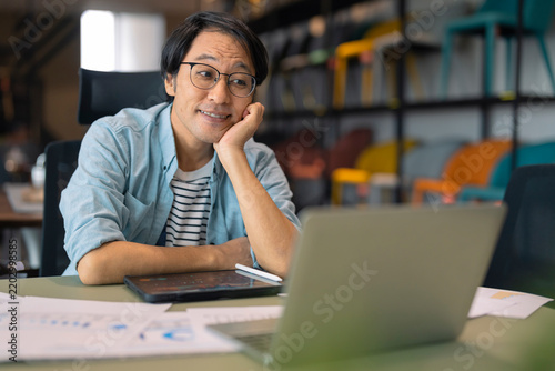 Принти на полотні Playful Asian businessman resting chin on hand at desk, daydreaming with thought