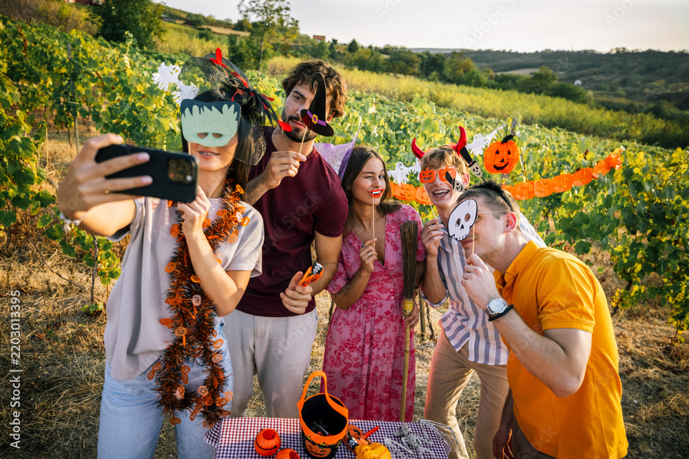 Obraz premium Group of friends in Halloween costumes holding paper masks and taking a selfie in a vineyard.