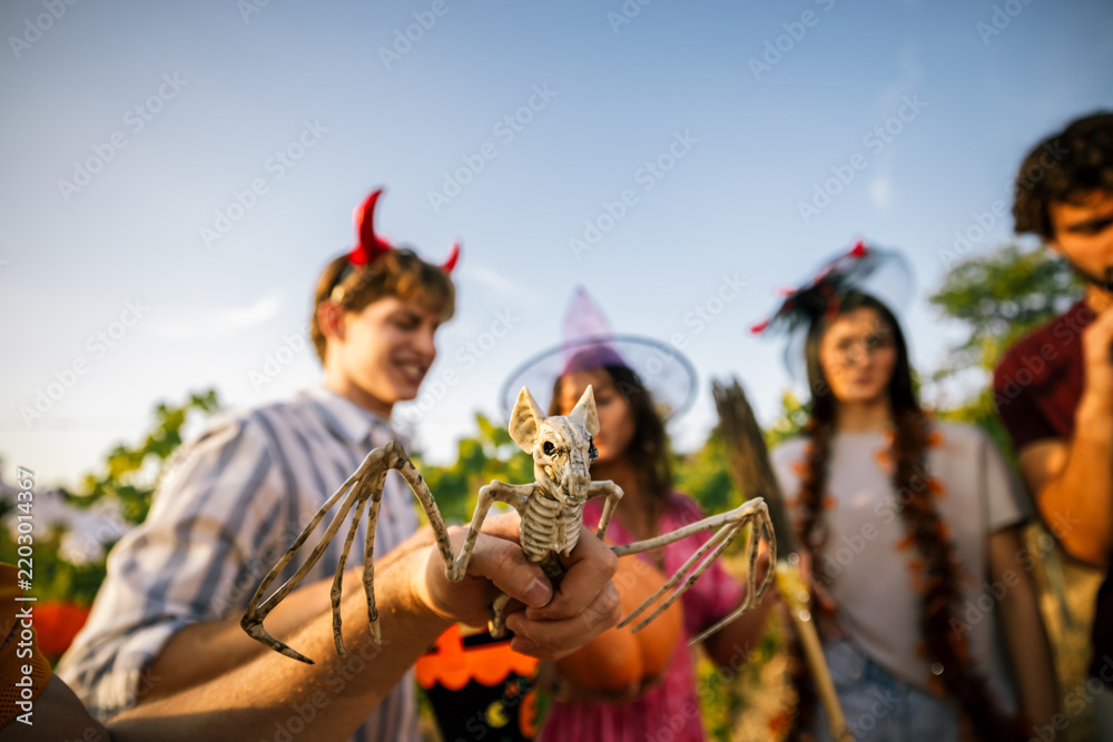 Obraz premium Close-up of a hand holding a skeleton bat prop with blurred friends in Halloween costumes.
