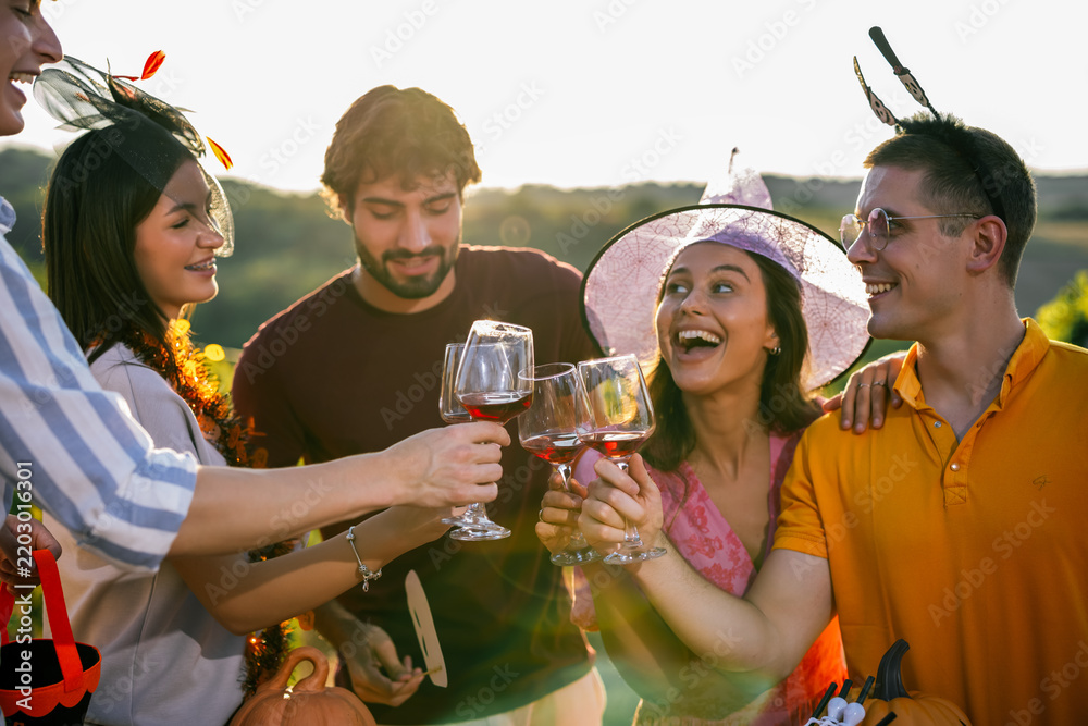Obraz premium Smiling friends in Halloween costumes laughing and making a toast with red wine at a sunny vineyard gathering.