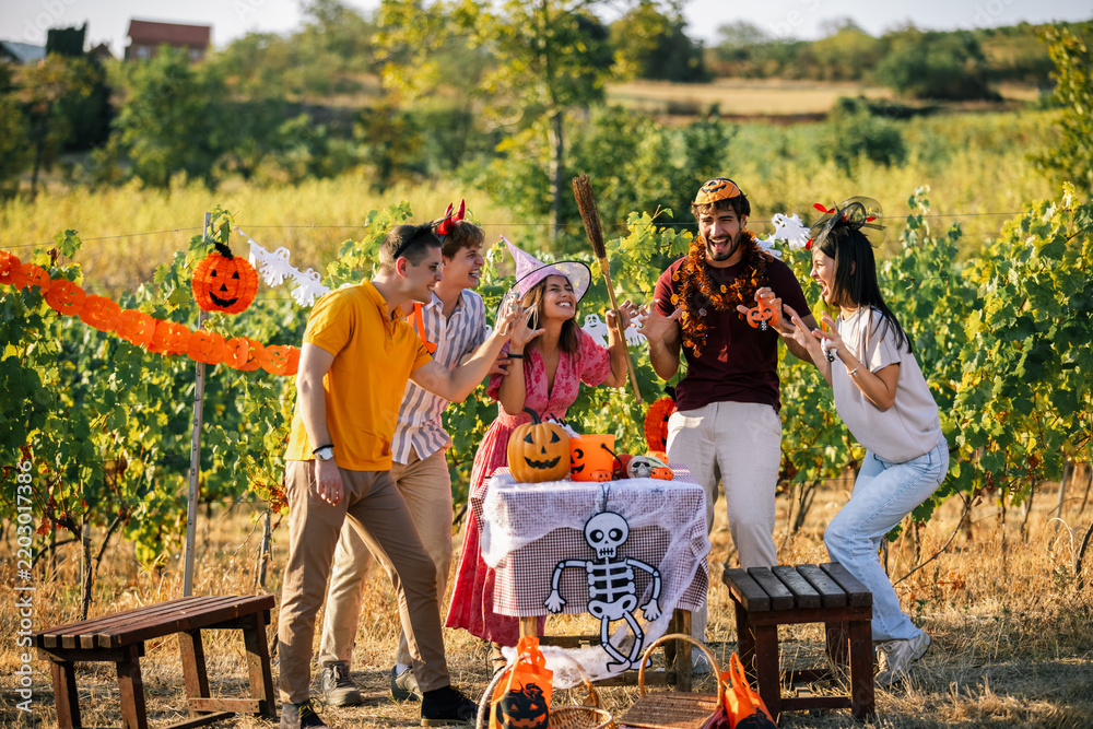 Obraz premium Five friends in Halloween costumes laugh and celebrate together beside a decorated table in a vineyard.