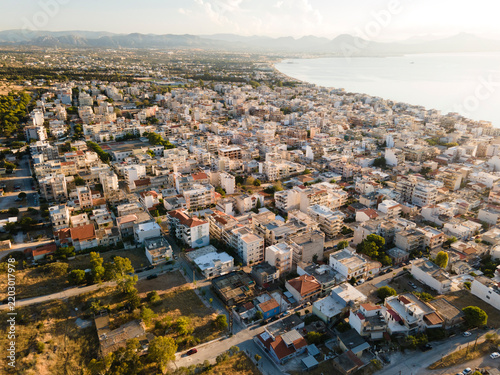 Aerial view over Loutraki, Greece, showing the town bathed in warm golden-hour sunlight, with the coastline and a stretch of beach visible in the distance. Peloponnese