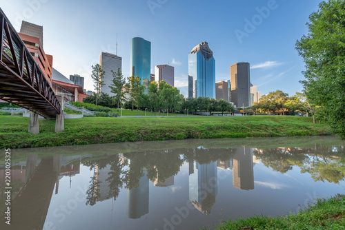 Houston, Texas skyline is reflected in the Buffalo Bayou. Prominent skyscrapers like the JPMorgan Chase Tower and Wells Fargo Plaza feature sleek, modern designs. 