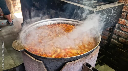 Hearty meat stew simmers vigorously in a cast iron cauldron with dense steam billowing outdoors. Authentic open fire cooking adds rich culinary texture to rustic food content.
