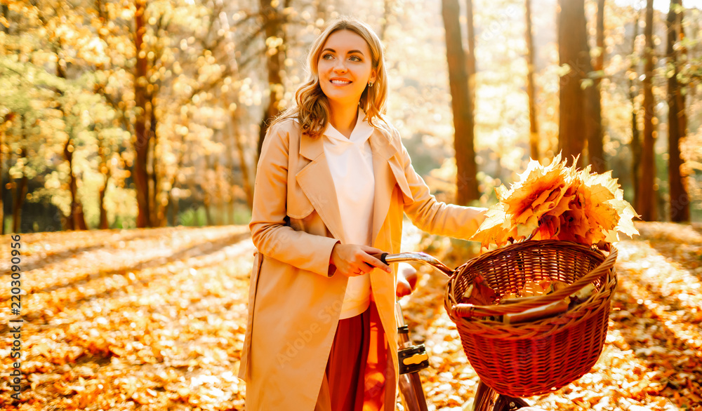 Obraz premium Woman in coat, holding bouquet of bright leaves, stands beside bicycle in an autumn forest. Smiling woman enjoys sunset while out for bike ride. Concept of active leisure, weekends.