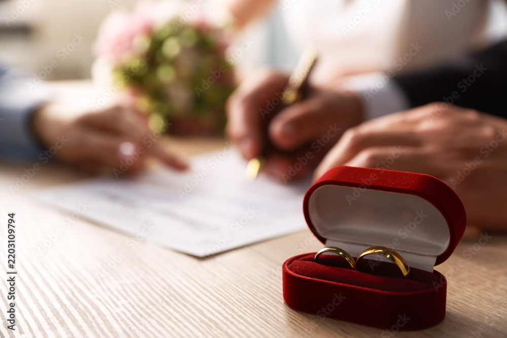 Obraz premium Notary helping couple with signing of marriage contract and wedding rings in box on wooden table indoors, selective focus