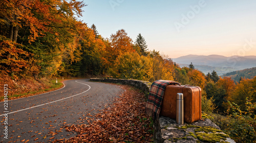 Vintage brown leather suitcase and metal thermos sitting on a stone wall beside a winding road during golden autumn season