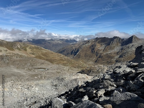 High-altitude panoramic mountain view in the Swiss Alps near Realp, Uri, showing jagged rocky peaks, dramatic ridges, alpine valleys and cloud layers under a deep blue sky in the wild landscape