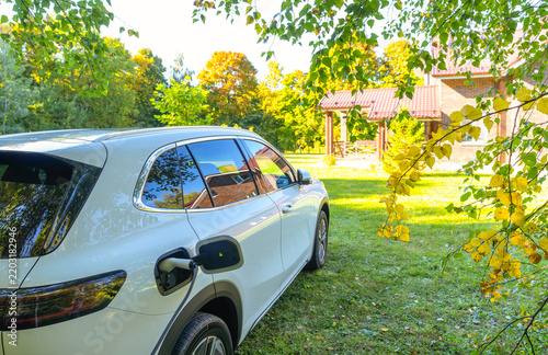 Electric vehicle charging process against the backdrop of a cozy country cottage and autumn foliage