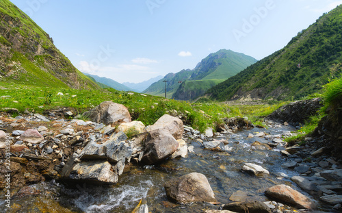 Panoramic view of a lush mountain gorge with a flowing stream, rocky terrain and cable cars crossing the slopes
