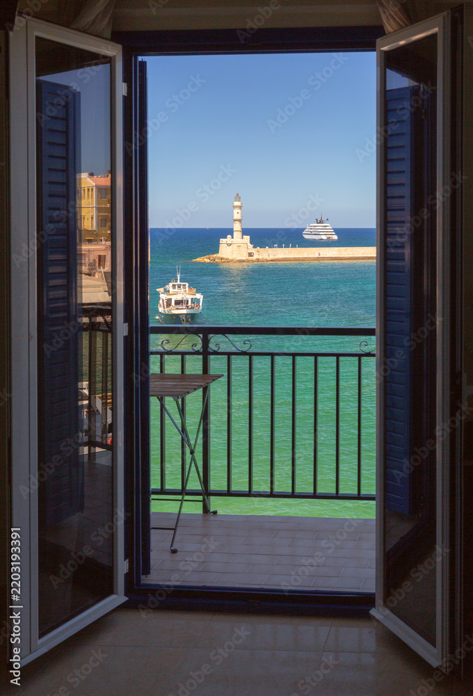 Fototapeta premium View through open balcony doors toward Chania Lighthouse, turquoise sea and boats in the Old Venetian Harbor of Chania, Crete, Greece.