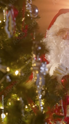 Close up video of Santa Claus with white beard and glasses hanging festive ornaments on decorated Christmas tree. Magical holiday scene with warm glowing lights and cozy atmosphere