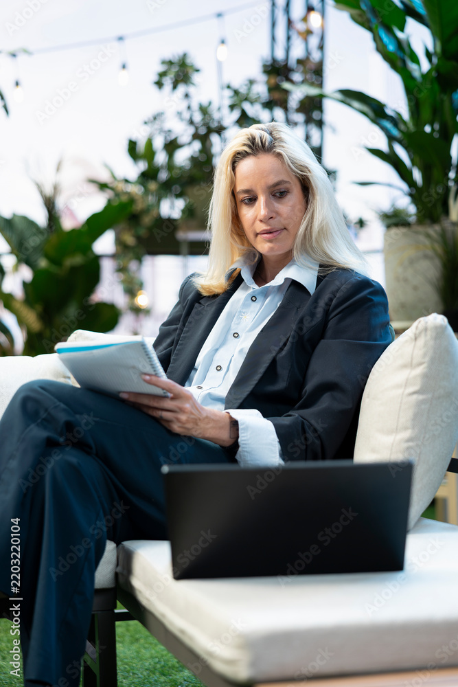Fototapeta premium Businesswoman reviews a notebook beside an open laptop in a bright rooftop workspace, writing notes while comparing and scheduling data on her agenda.