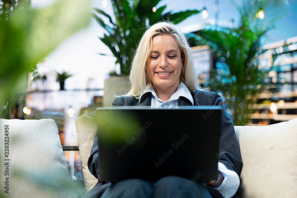 Fototapeta premium Professional businesswoman types on a laptop from a cushioned rooftop lounge surrounded by greenery, illustrating concentrated remote work in a comfortable urban outdoor workspace.