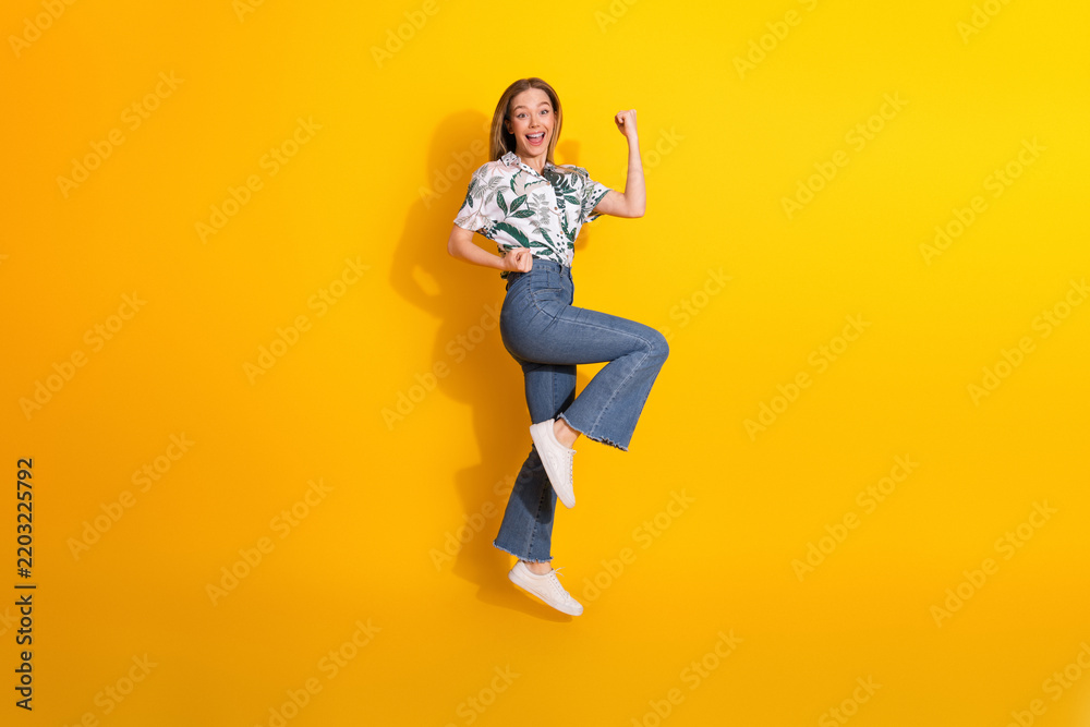 Fototapeta premium Cheerful young woman in fashionable casual attire jumping with joy on yellow background showcasing vibrant lifestyle