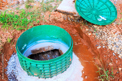 Septic tank plastic sewer access cover is open in dug area with soil, gravel nearby.