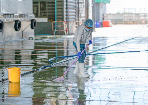 Worker wearing protective gear cleaning outdoor concrete floor with broom and water hose near industrial building.
