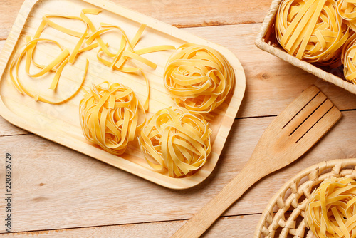 Nests of dry, uncooked tagliatelle arranged on a wooden tray, accompanied by loose pasta strands and baskets on a rustic table.