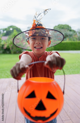 Asian boy in witch hat holding out pumpkin bucket for trick or treat