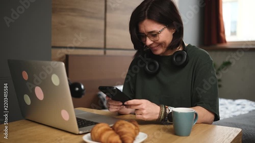 Woman taps her smartphone with both hands at the kitchen table, enjoying coffee and a croissant