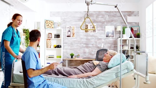 Two nurses consult an older patient in bed while one takes notes, showing coordinated care, clinical teamwork, communication and senior health support.