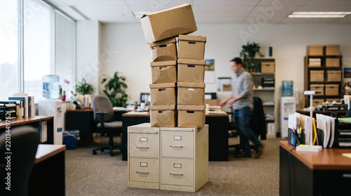 Unstable stack of cardboard archive boxes on a filing cabinet in a busy office