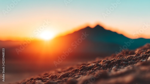 A vibrant sunrise illuminates a mountain silhouette with warm orange and blue tones, highlighting textured rocky foreground in a serene natural landscape.