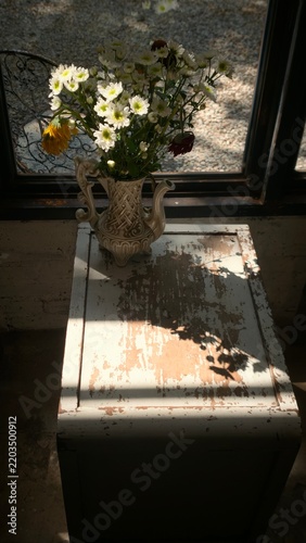 A bouquet of white chrysanthemums arranged in an ornate ceramic vase on a rustic wooden table, illuminated by warm natural sunlight through a window, creating a cozy vintage atmosphere.