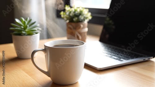 Cinematic close-up of a steaming cup of hot coffee beside a laptop on a wooden desk. Warm natural sunlight creates a cozy home office atmosphere, ideal for remote work and morning routines.