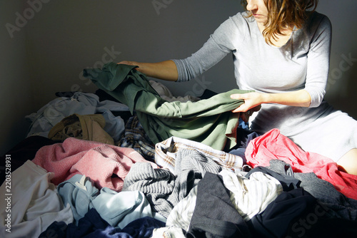 Картината върху платно Woman seated amid massive heap of mixed clothes on bed, holding green garment with face turned downward in focused assessment