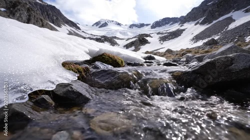 Crystal-clear mountain stream cascades over ancient, worn rocks, winding through a pristine snow-covered alpine landscape towards distant majestic peaks under a vast sky
