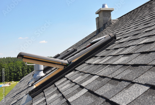 Skylight on asphalt shingle house roof with chimney and ventilation pipe.