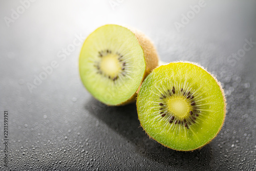 Two sliced fresh kiwi halves with brown fuzzy skin and green flesh, placed on a dark surface with water droplets. Close-up view of ripe fruit.