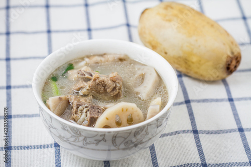 Фотография A bowl of simmered lotus root and pork ribs soup, with a whole raw lotus root beside it on a checkered tablecloth