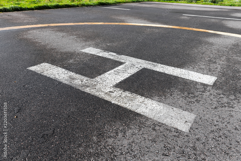 Obraz premium Helipad landing marking painted on asphalt tarmac showing a weathered white H and faded yellow circle, conveying aviation, emergency readiness, texture and urban outdoor infrastructure